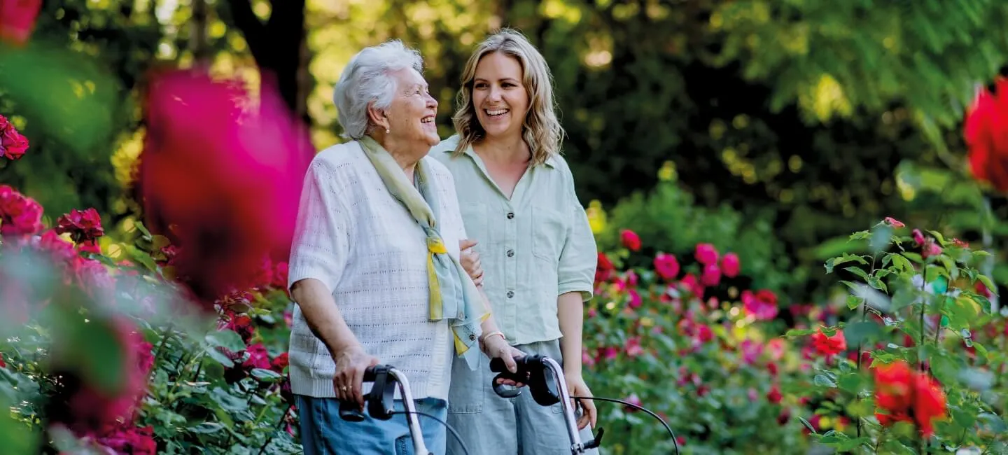 Pflegerin begleitet eine Seniorin mit Rollator durch einen Rosengarten